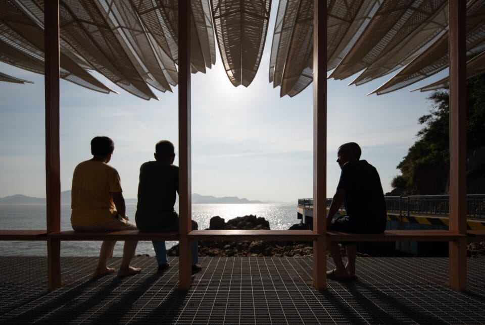 the view underneath a seaside pavilion made of white feather-like blades. three people sit on a bench and look out