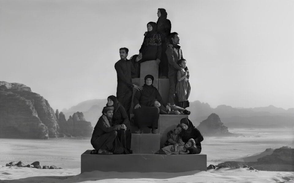 a black-and-white photograph in the Jordanian desert of a group of people in black garments perched on a stack of boxes to create a monolith-like form