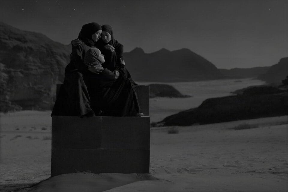 a black-and-white photograph in the Jordanian desert of a group of people in black garments perched on a stack of boxes to create a monolith-like form