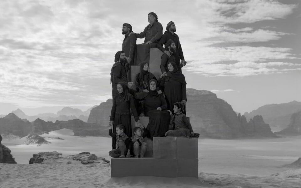 a black-and-white photograph in the Jordanian desert of a group of people in black garments perched on a stack of boxes to create a monolith-like form