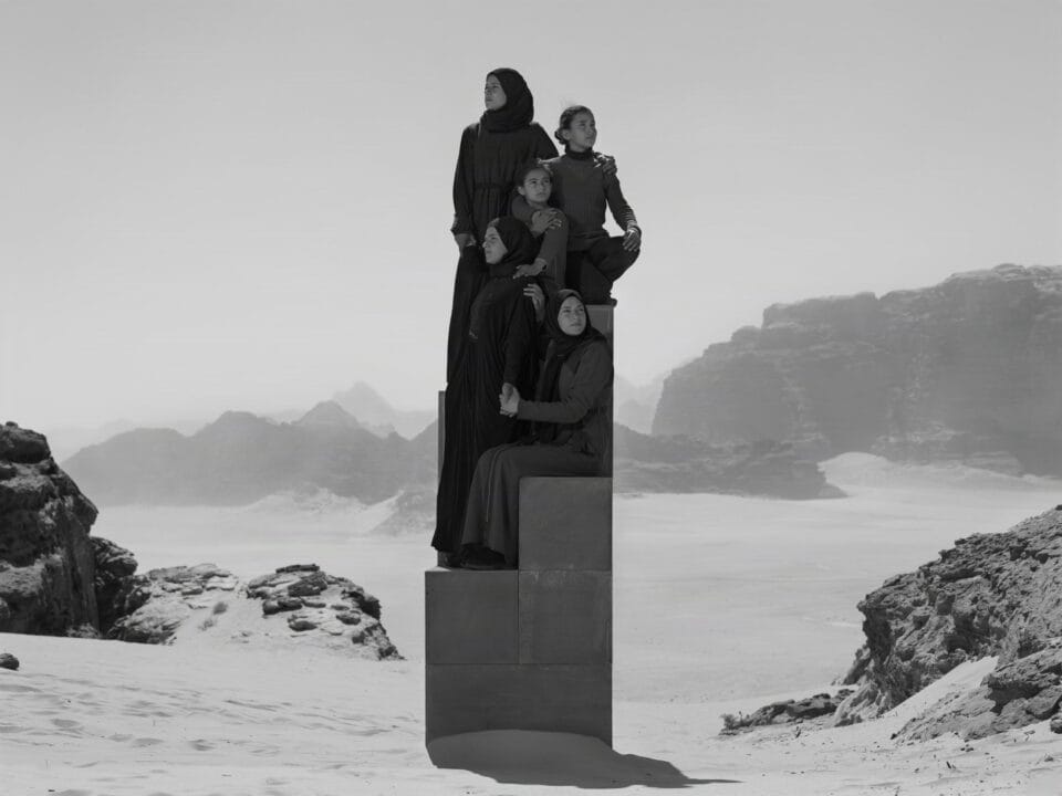 a black-and-white photograph in the Jordanian desert of a group of people in black garments perched on a stack of boxes to create a monolith-like form