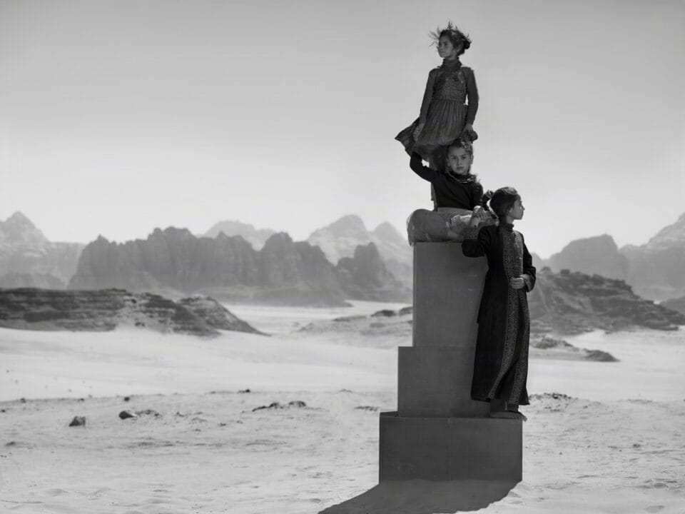 a black-and-white photograph in the Jordanian desert of two children in black garments perched on a stack of boxes to create a monolith-like form