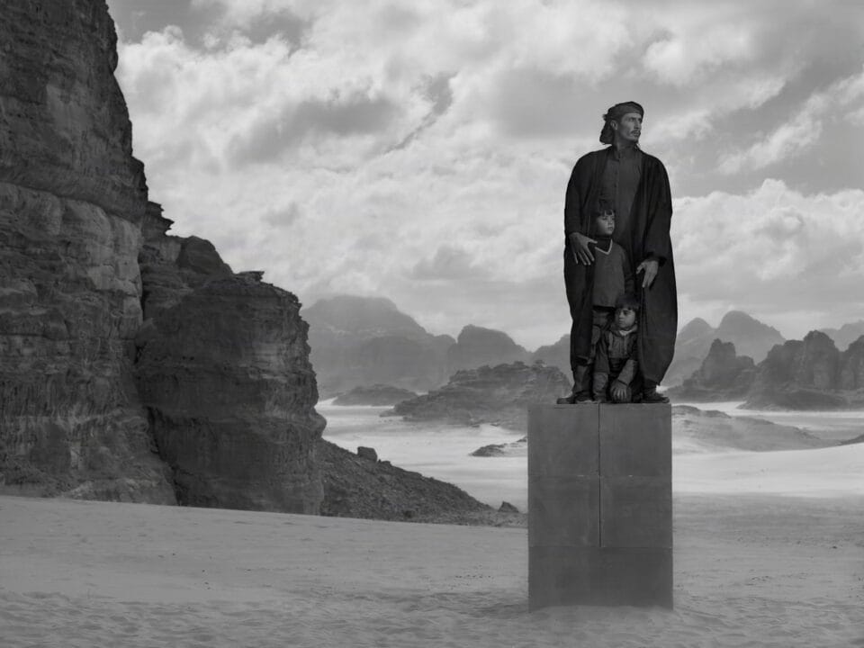 a black-and-white photograph in the Jordanian desert of two people in black garments perched on a stack of boxes to create a monolith-like form