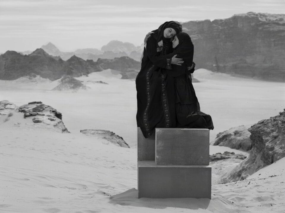a black-and-white photograph in the Jordanian desert of two children in black garments perched on a stack of boxes to create a monolith-like form