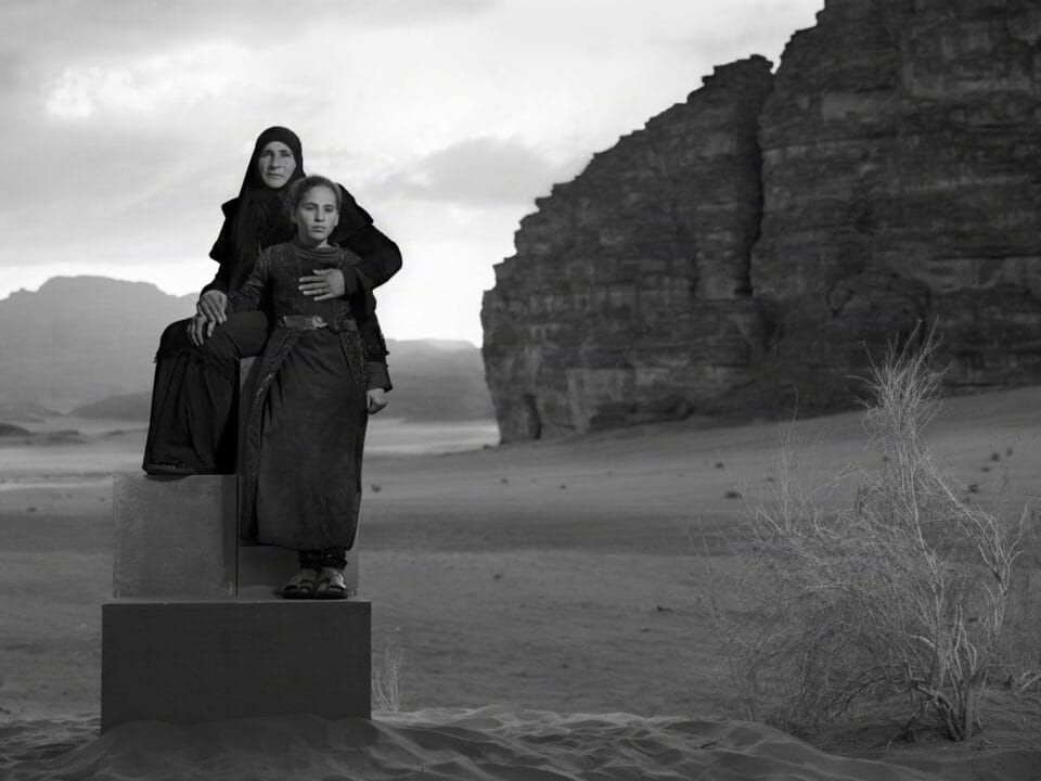 a black-and-white photograph in the Jordanian desert of two people in black garments perched on a stack of boxes to create a monolith-like form