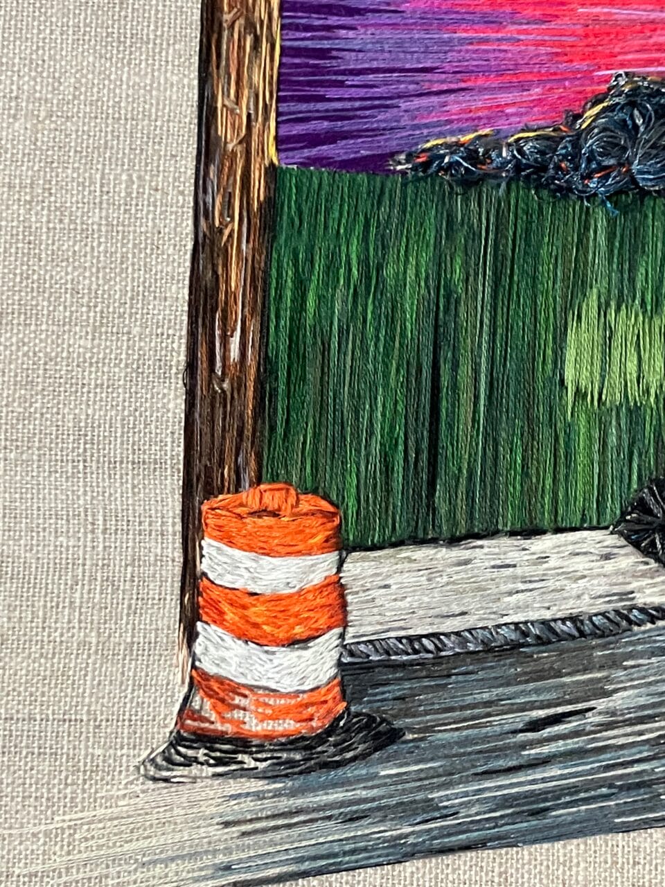 detail of a vibrant sunrise peeks over a green fence. an orange construction barrel sits on the sidewalk in front