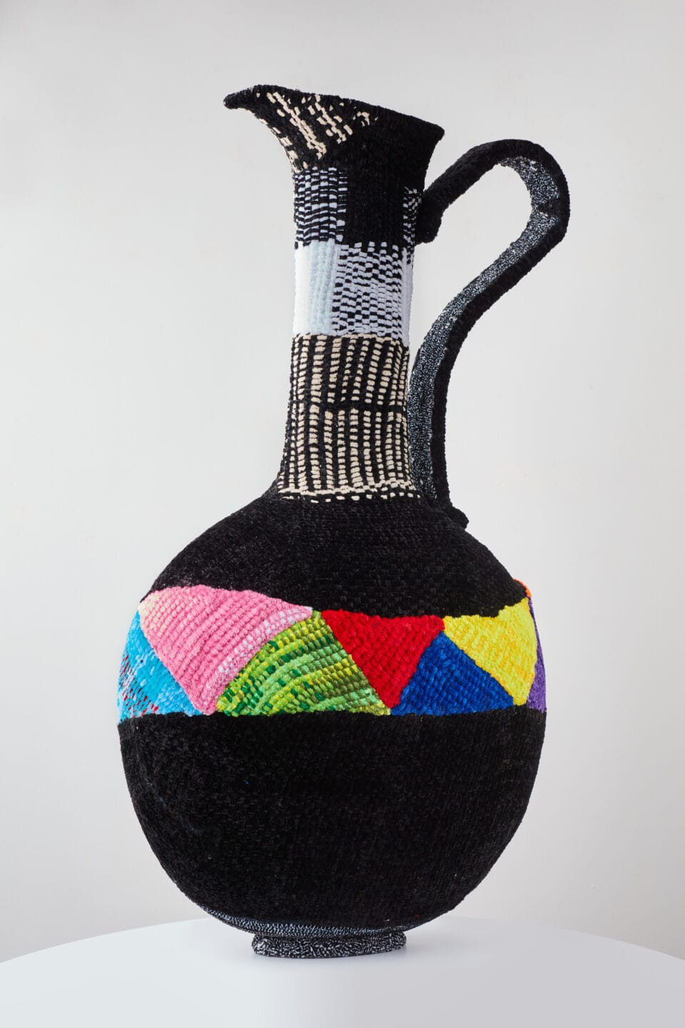 a tall vessel made from woven pipe cleaners in shades of black and gray with geometric colorful detail