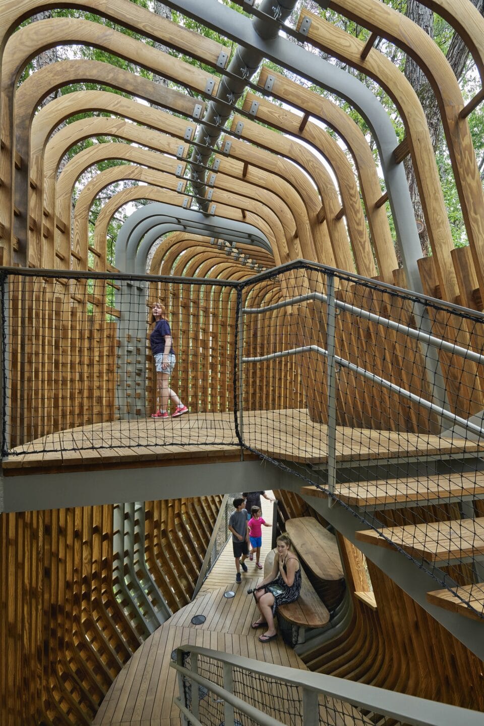 the interior of a contemporary, open-plan tree house in a public forest, featuring numerous stairwells and passages, surrounded by trees, with people inside for scale