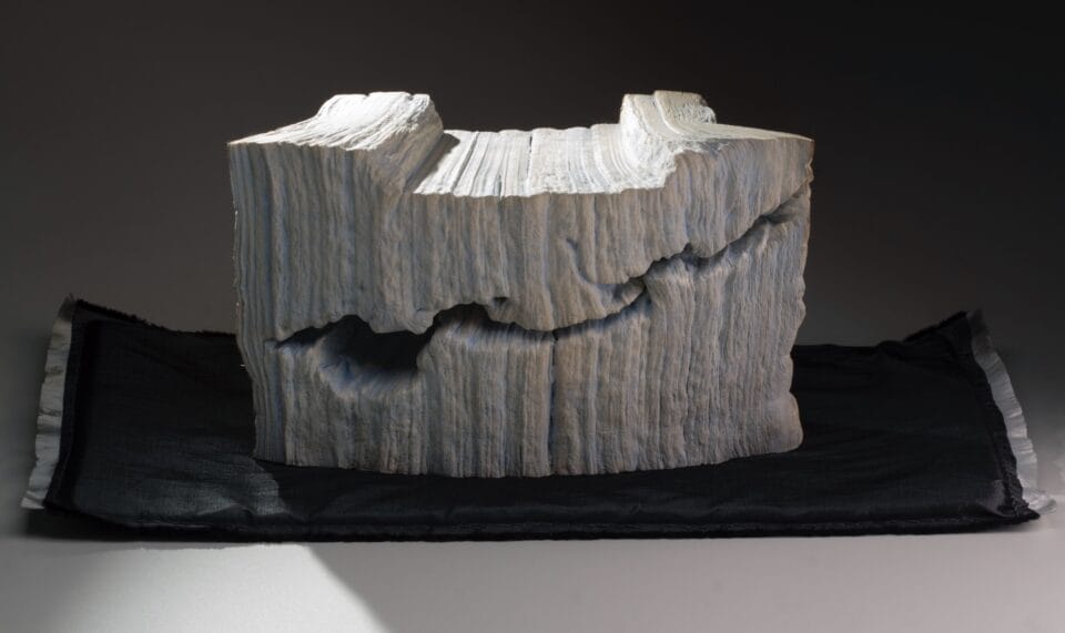a sculpture made from a set of large books, covers removed to reveal the stitched binding, which has been water-blasted to soften and manipulate the pages into a form resembling a large mountain or glacier with a large crevasse