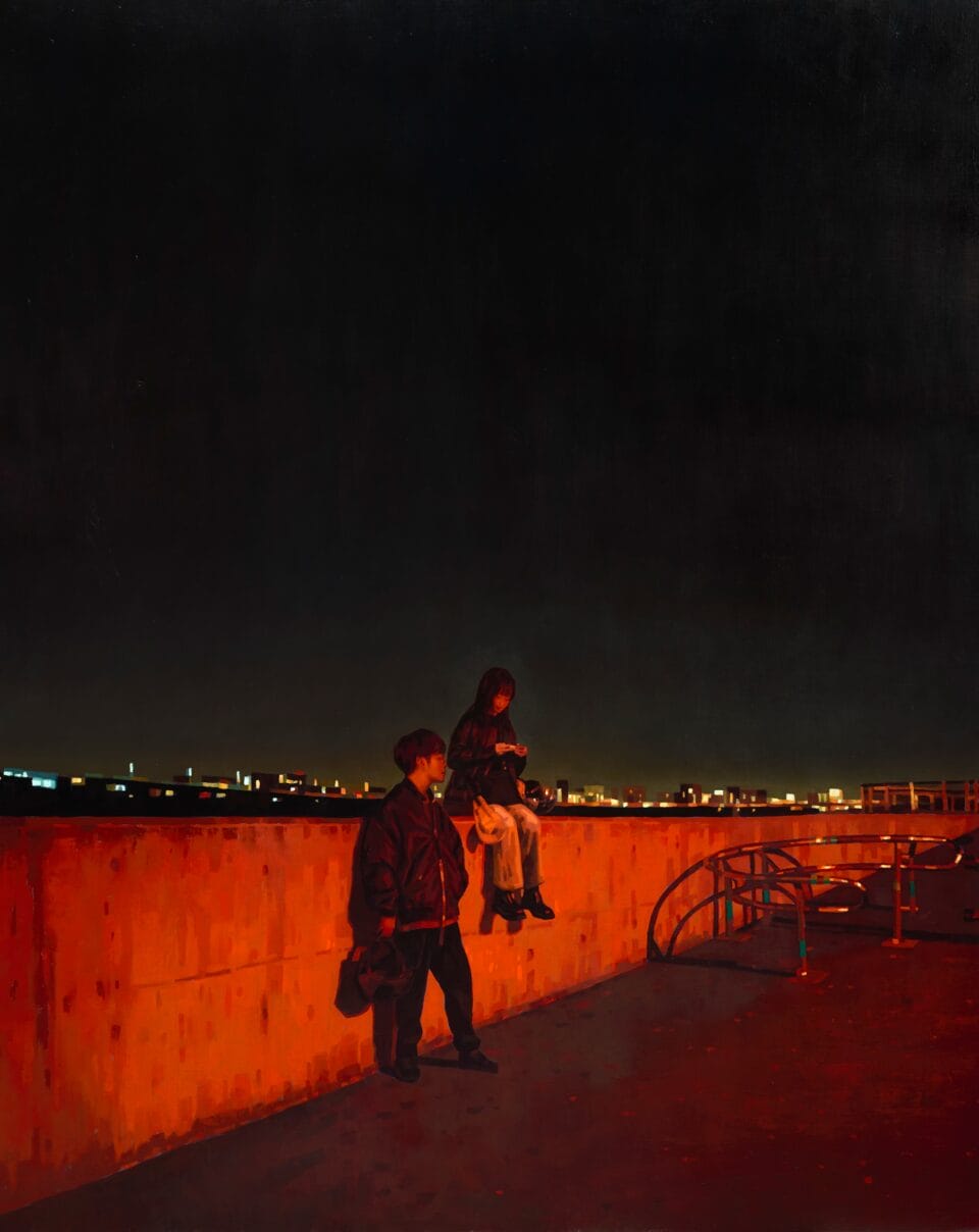 one person sits on a concrete wall while another stands nearby. both are illuminated by a deep orange light