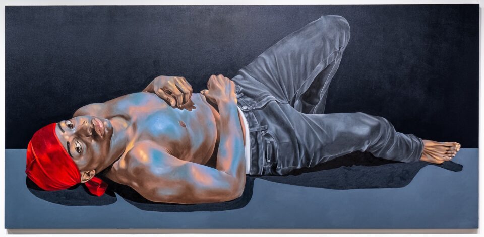 a wide horizontal portrait of a Black man wearing a red durag, no shirt, and jeans, lies on the ground and looks over at the viewer