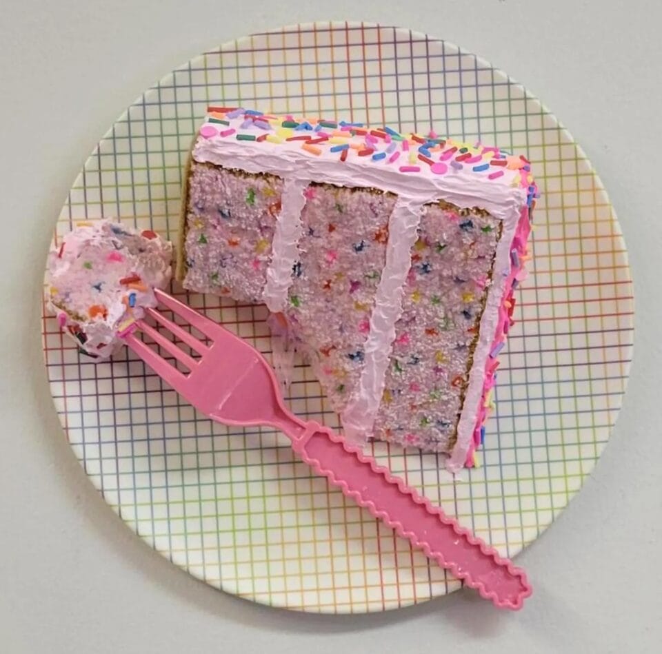 a polymer clay sculpture of a piece of layer cake with embroidered panels for the sponge and a pink plastic fork with a bite of cake on the end, sitting on a retro plate