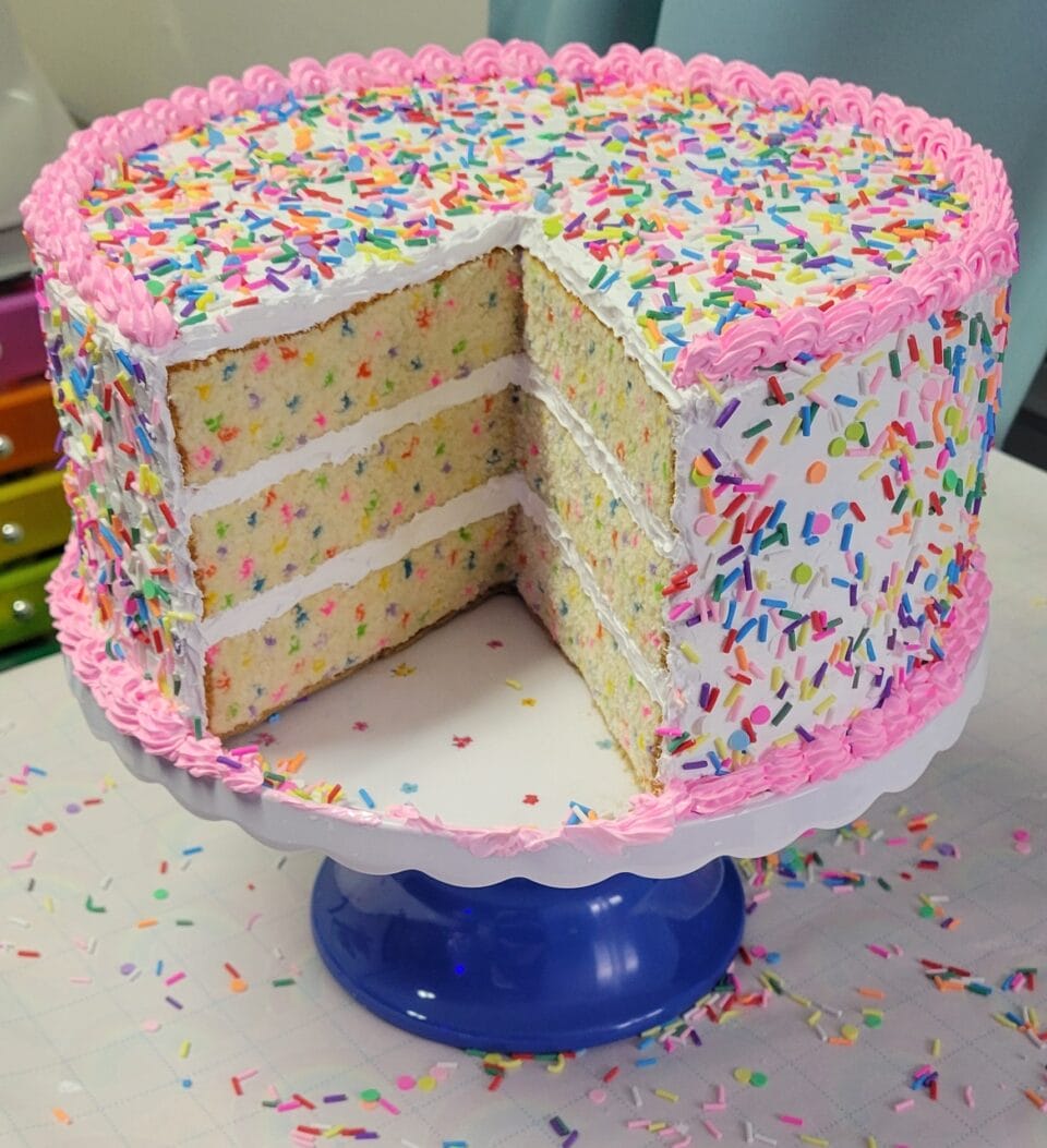 a small sculpture of a layer cake, with realistic frosting details, sprinkles, and embroidery on that looks like the texture of fluffy sponge where a slice has been removed