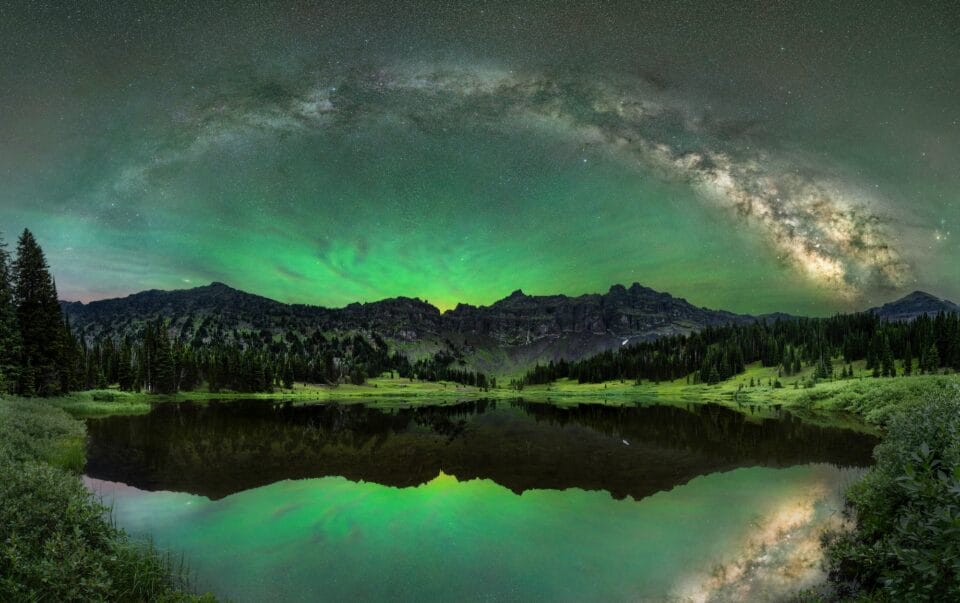 the Milky Way expands in an arc across a watery, mountainous landscape illuminated by green aurora borealis