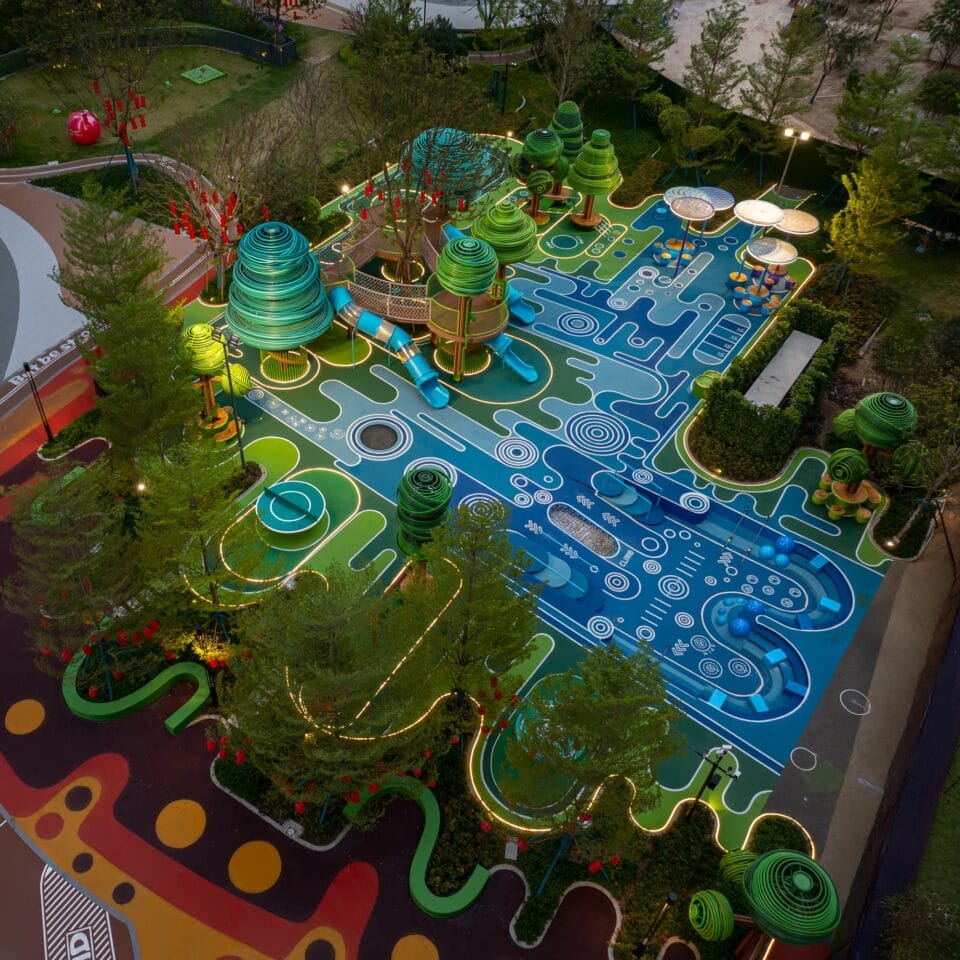 an aerial view at dusk of an elaborately designed playground with blue and green designs on the ground and wire-like sculptural trees holding up bridges and play equipment