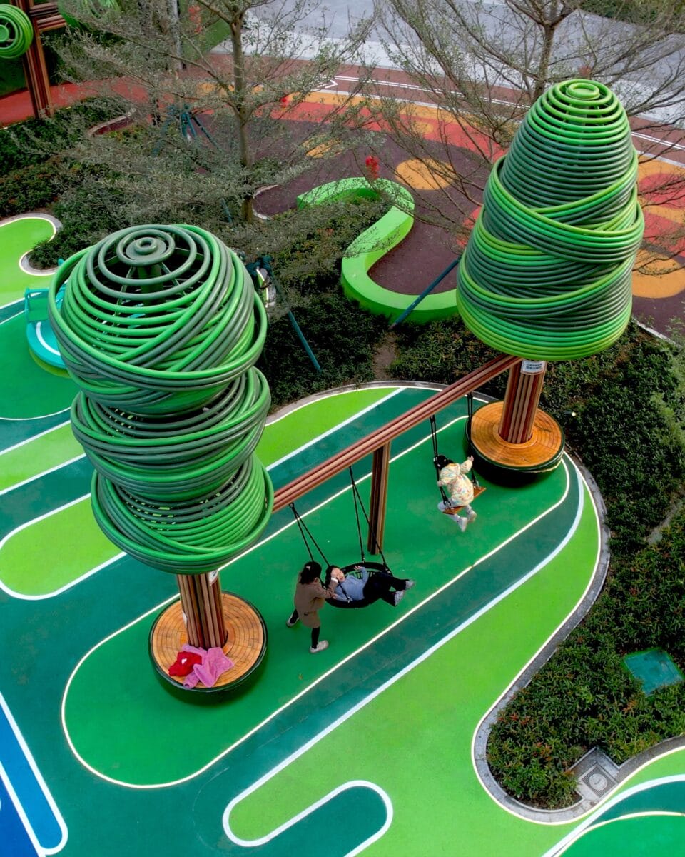 an aerial view of part of an elaborately designed playground with blue and green designs on the ground and wire-like sculptural trees holding up bridges and play equipment