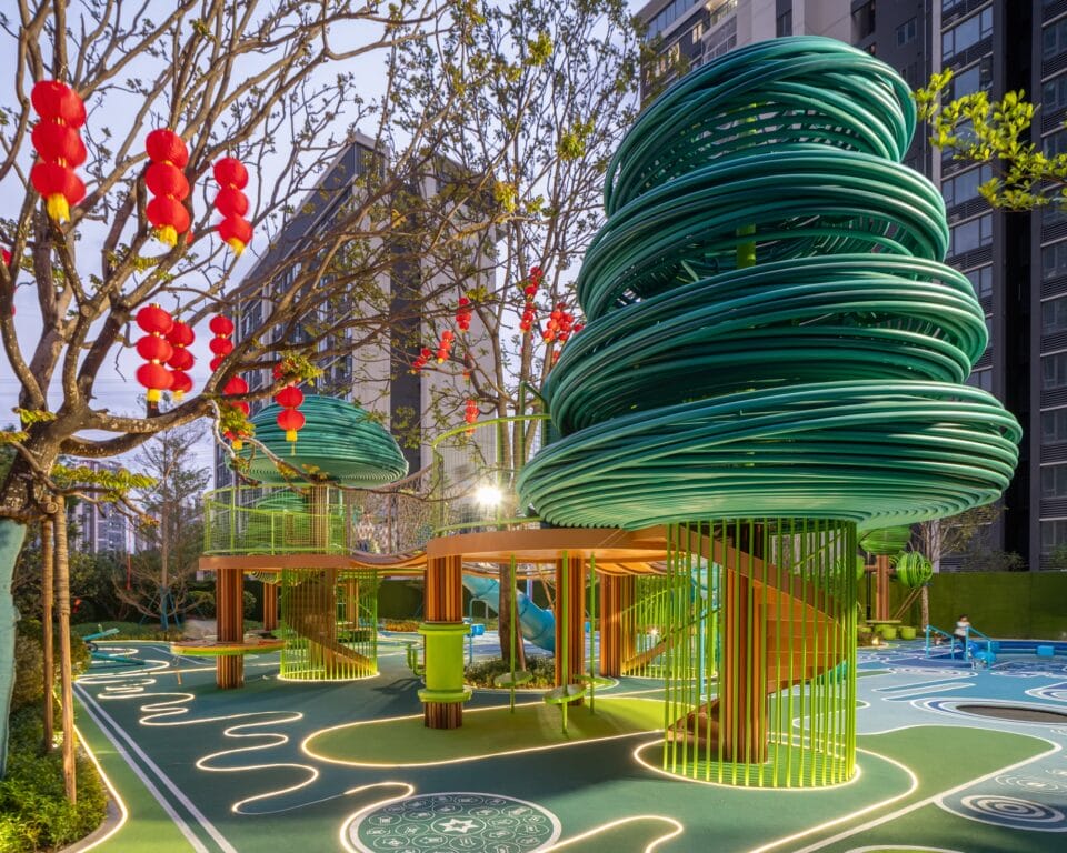 an evening view of an elaborately designed playground with blue and green designs on the ground and wire-like sculptural trees holding up bridges and play equipment