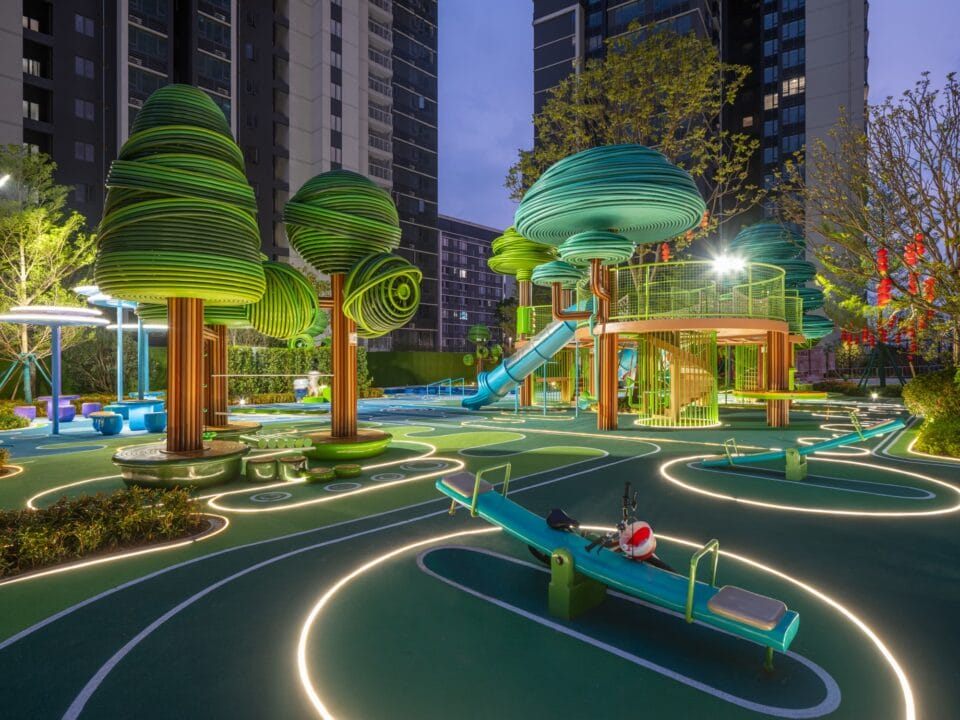 a nighttime view of an elaborately designed playground with blue and green designs on the ground and wire-like sculptural trees holding up bridges and play equipment