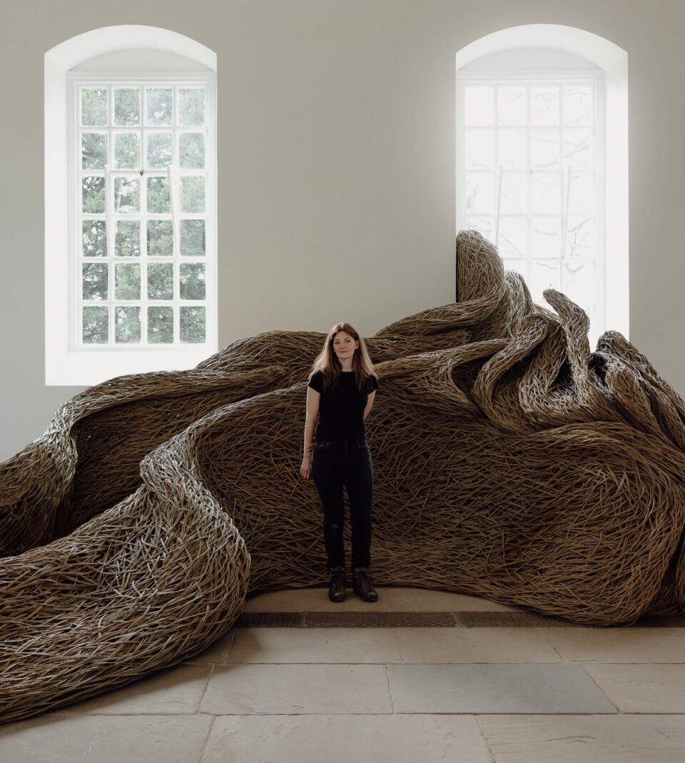 Artist Laura Ellen Bacon standing in front of a sculptural installation that moves through an architectural space and interacts with windows, made of woven willow branches