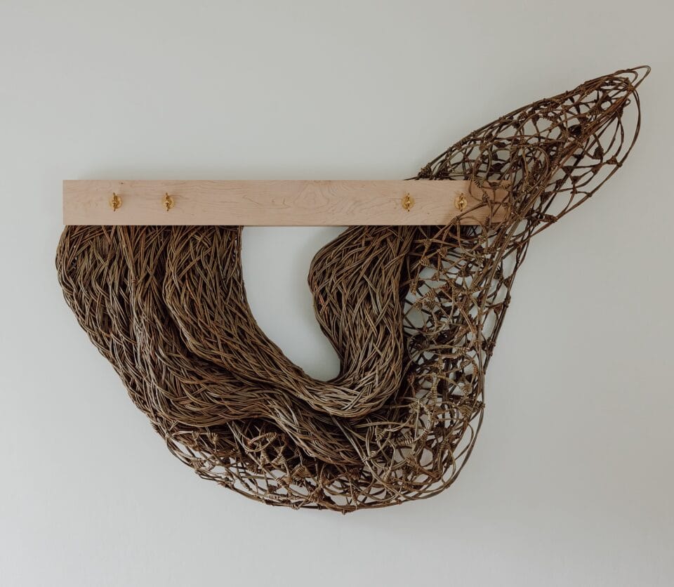a sculpture made from willow, installed on the wall, featuring an organic, woven form that ineracts with a horizontal board