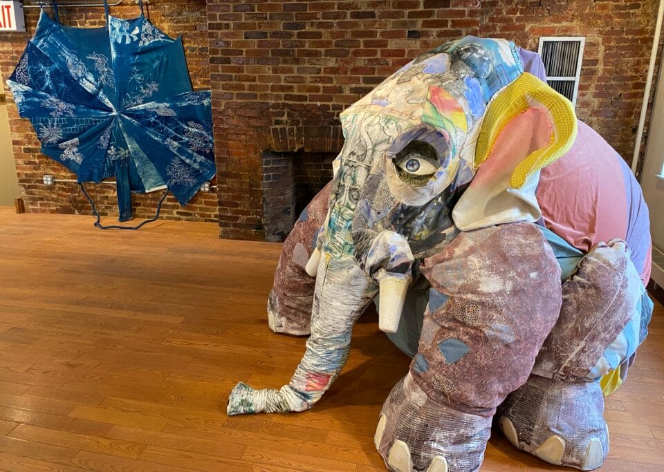 a patchwork elephant sculpture on the right with an umbrella like blue work suspended in the background