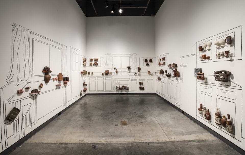 a large, room-size installation in a gallery featuring line drawings on the walls of a kitchen with three-dimensional reliefs and sculptures in ceramic of vessels and other objects attached to the wall