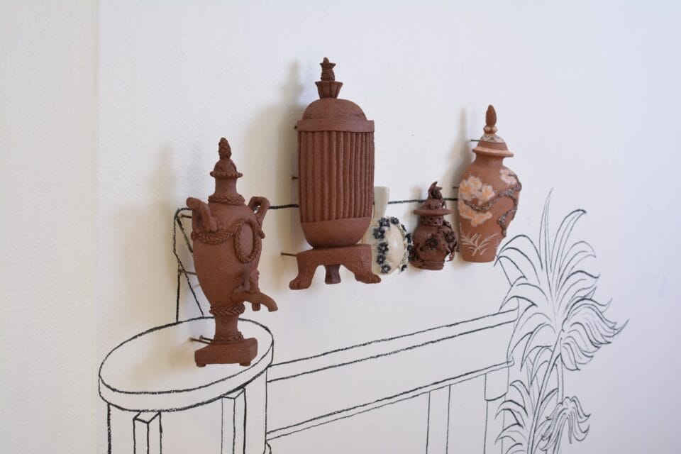 a group of ceramic sculptural reliefs of vases, decorative vessels, and other objects on a wall, against a line drawing of a mantle in a gallery space