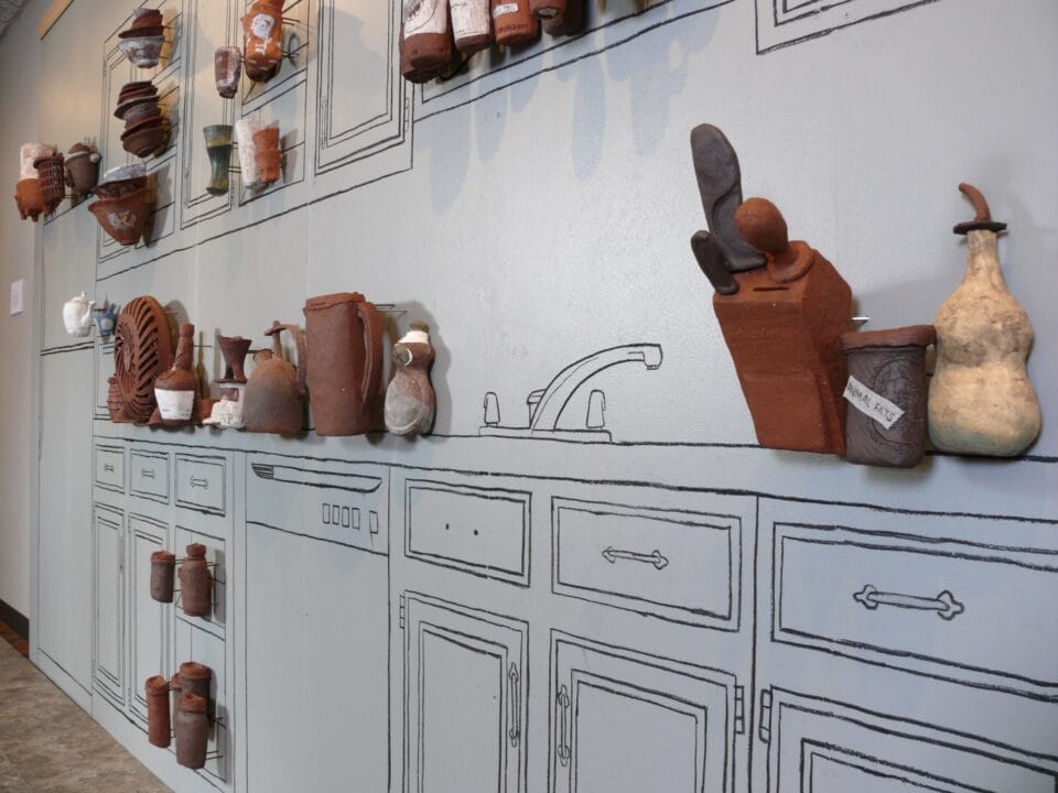 a detail of a gallery wall outlined with kitchen cabinets and counters, with three-dimensional reliefs of ceramic vessels and objects installed on the wall