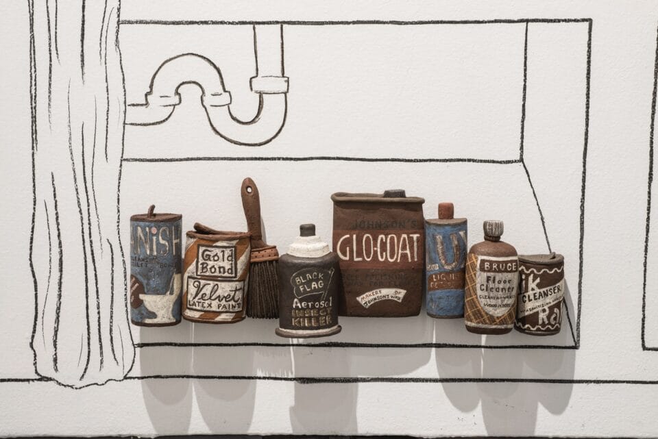 a group of ceramic sculptural reliefs of food, vessels, cups, and other objects on a wall, against a line drawing of a kitchen cabinet