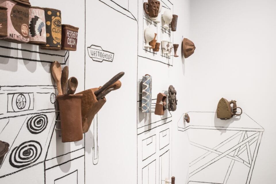 a detail of a large, room-size installation in a gallery featuring line drawings on the walls of a kitchen with three-dimensional reliefs and sculptures in ceramic of vessels and other objects attached to the wall