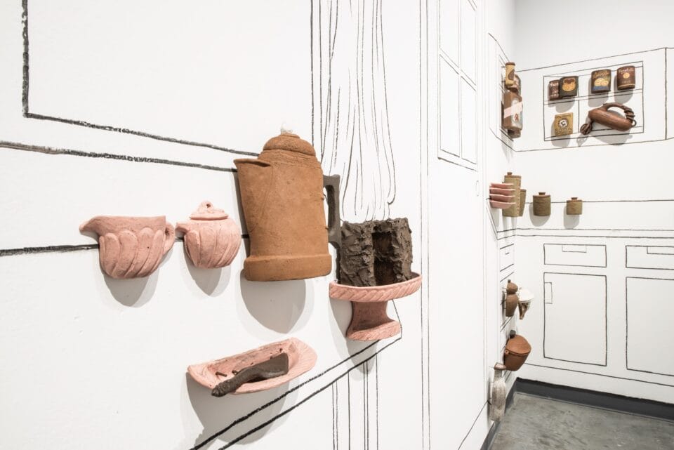 a group of ceramic sculptural reliefs of food, vessels, cups, and other objects on a wall, against a line drawing of a kitchen
