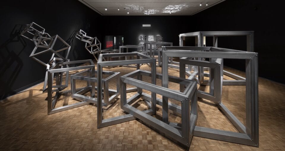 a museum installation of large cubic metal frames interlocked in a giant jumble, inside of a room painted black