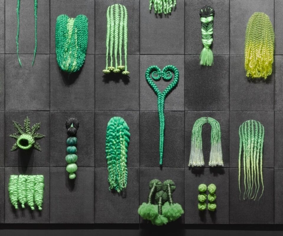 a detail of an art installation of numerous green wigs in a variety of braided styles on a black wall