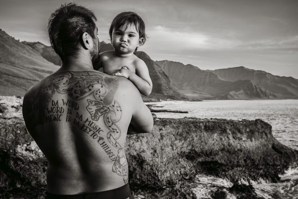 a black-and-white photograph of a man with tattoos on his back holding his daughter in front of a coastal landscape