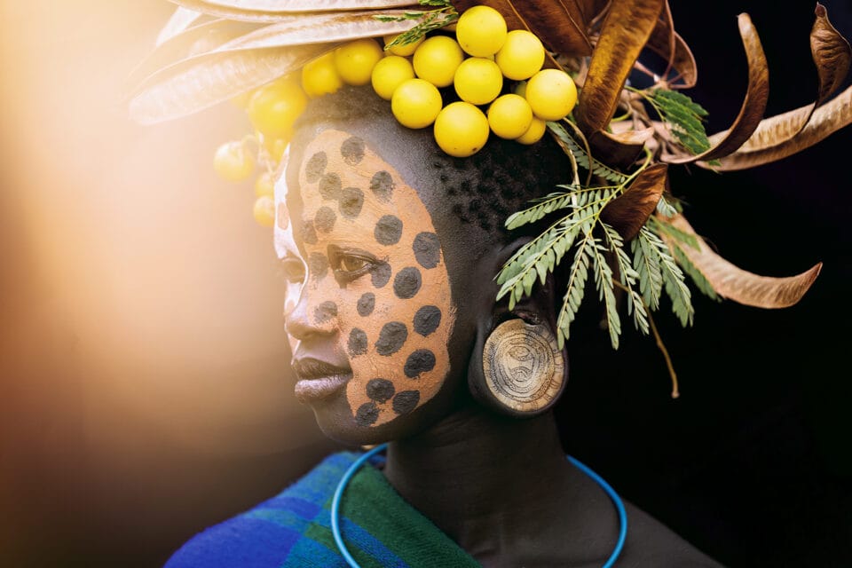 a portrait of a young Black woman with black-and-orange face paint and an elaborate headdress of yellow spheres, twigs, and other natural objects