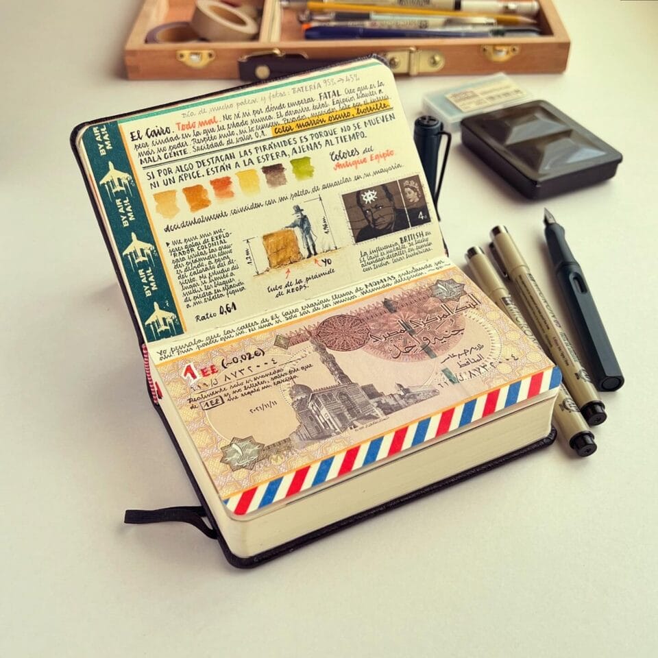 a small sketchbook with elaborately designed travel notes and drawings, on a table with artmaking tools like pens and ink