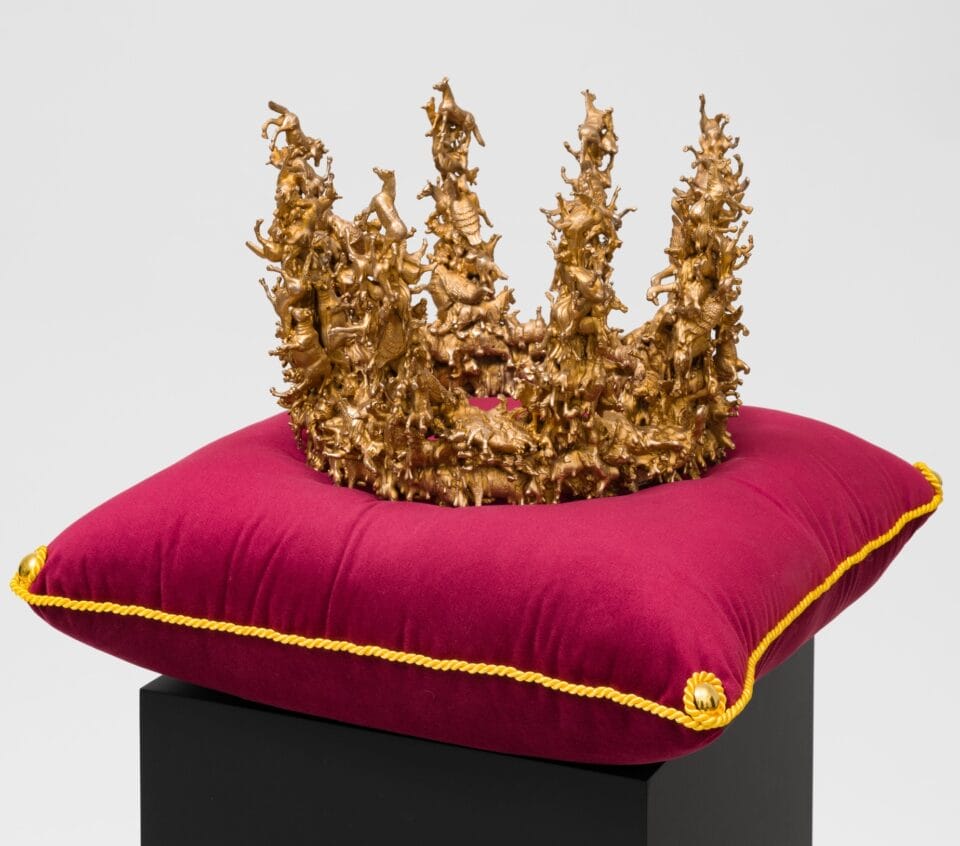 a sculpture of a bronze crown composed of tiny birds and other animals, coated in metallic silver, situated on a red velvet cushion