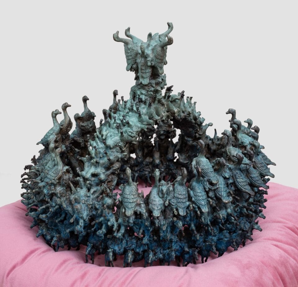 a sculpture of a patinated bronze crown composed of tiny birds and other animals, situated on a pink velvet cushion
