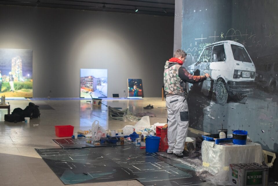 a young man works on a painting inside of a museum, with materials and other paintings nearby