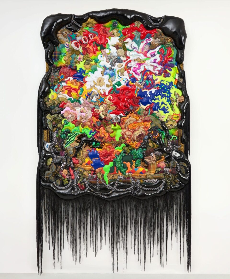 a wall-mounted abstract textile artwork with a shiny black border and long black fringe at the bottom, with colorful, almost floral quilted patterns in the center