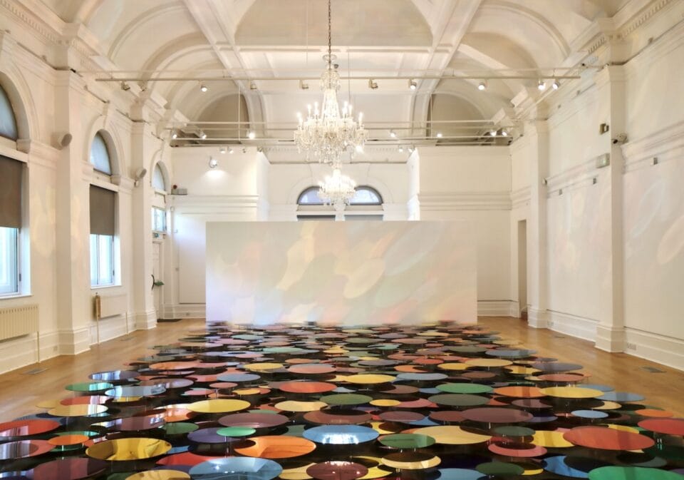 a floor installation in a white-walled gallery space of numerous shiny, colorful circles