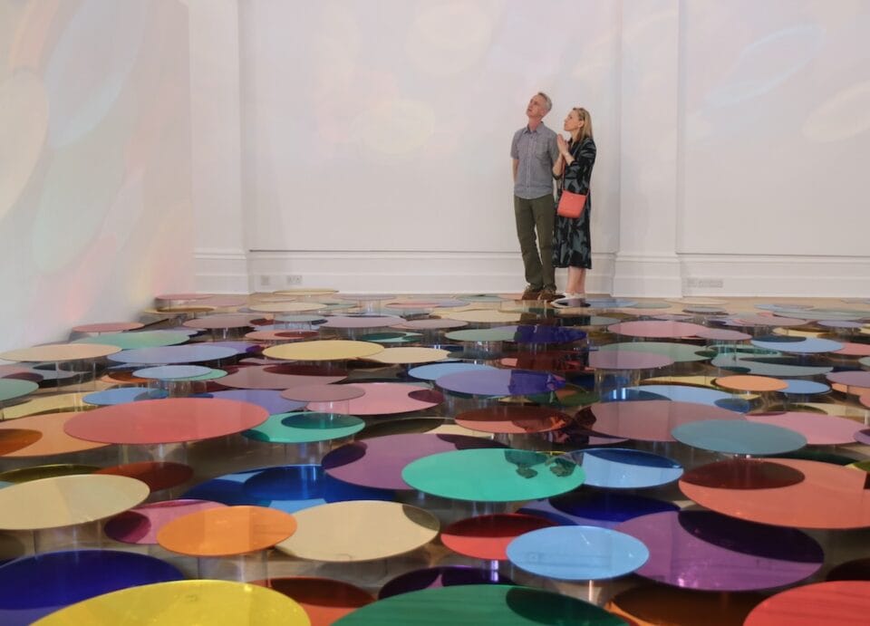 two people stand inside of a white gallery space, alongside a floor installation of numerous shiny, colorful circles